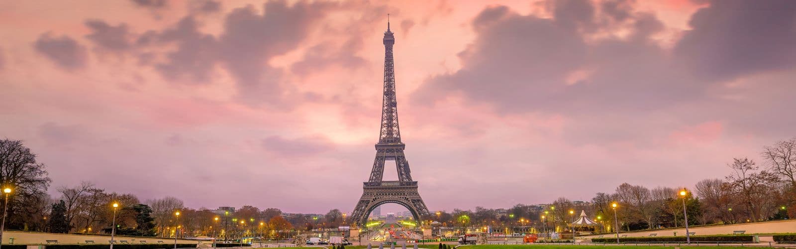 The Eiffel Tower stands front and center, surrounded by the lights of the city of Paris at sunset. 